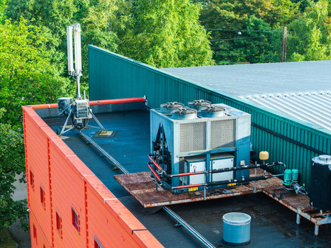 Aerial view of an industrial rooftop featuring a large HVAC chiller unit and a cell tower antenna set against a backdrop of lush green trees in Riga Latvia. Aerial view of an industrial rooftop featuring a large HVAC chiller unit and a cell tower antenna set against a backdrop of lush green trees in Riga Latvia.