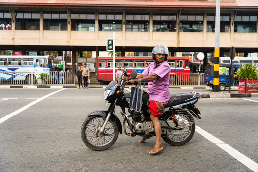 Sri Lanka 07 January 2026 View of a man wearing a helmet and sitting on a motorcycle at a city intersection with a large building and buses in the background.