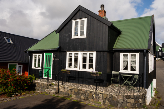 View of a traditional black wooden house with a green corrugated metal roof a bright green door and white framed windows situated on a stone foundation in the Faroe Islands. View of a traditional black wooden house with a green corrugated metal roof a bright green door and white framed windows situated on a stone foundation in the Faroe Islands.