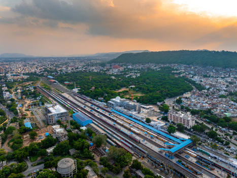 Aerial view of Udaipur Railway Station with tracks and trains surrounded by urban buildings lush trees and distant hills under a warm golden hour sky in Udaipur Rajasthan India. Aerial view of Udaipur Railway Station with tracks and trains surrounded by urban buildings lush trees and distant hills under a warm golden hour sky in Udaipur Rajasthan India.
