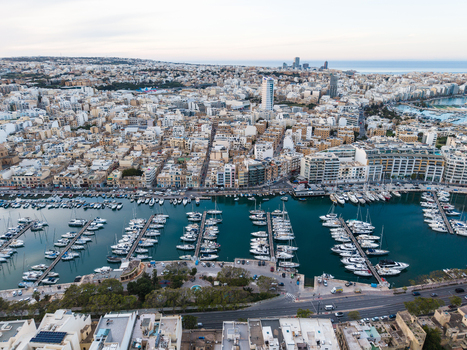 Aerial view of a bustling marina filled with white yachts and sailboats alongside dense urban architecture and coastal buildings in Malta. Aerial view of a bustling marina filled with white yachts and sailboats alongside dense urban architecture and coastal buildings in Malta.