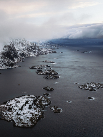 Aerial view of a dramatic coastline where snow dusted islands meet the dark swirling sea under a brooding sky Henningsv&aelig;r Nordland Norway.