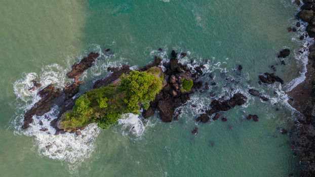 Aerial view of a rocky islet covered in lush green trees surrounded by turquoise ocean waves and coastal rocks in Ayah Central Java Indonesia. Aerial view of a rocky islet covered in lush green trees surrounded by turquoise ocean waves and coastal rocks in Ayah Central Java Indonesia.