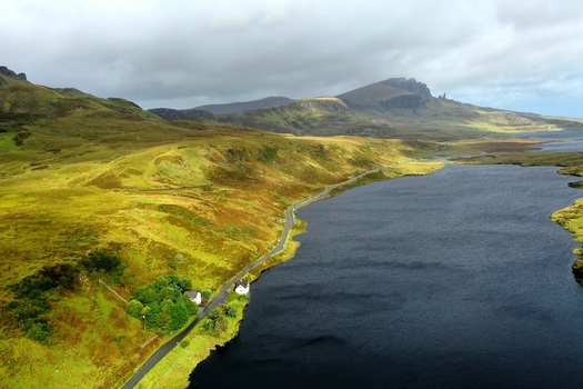 Aerial view of The Storr rising above rolling green hills and a winding road along the shore of a dark lake in Portree Scotland United Kingdom.