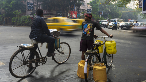 View of two men with bicycles and yellow containers pause at a bustling intersection with a blur of traffic Kolkata West Bengal India. View of two men with bicycles and yellow containers pause at a bustling intersection with a blur of traffic Kolkata West Bengal India.