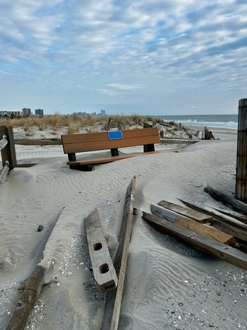 View of Ventnor City Beach featuring a wooden bench partially buried in sand dunes with scattered wooden debris and the ocean under a cloudy sky Ventnor City New Jersey United States.