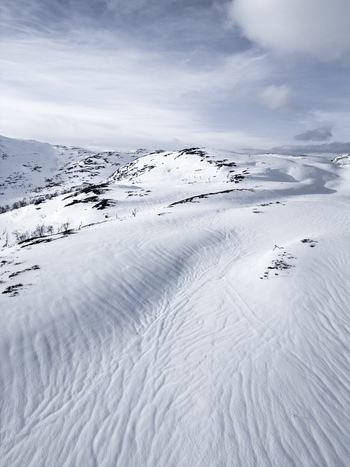 Aerial view of snow covered mountain slopes with wind blown textures and rolling hills under a cloudy winter sky in Kvaloya Troms Norway. Aerial view of snow covered mountain slopes with wind blown textures and rolling hills under a cloudy winter sky in Kvaloya Troms Norway.