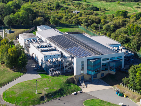 Aerial view of a modern data center facility featuring rooftop solar panels and industrial cooling units surrounded by lush green landscape in the United Kingdom.