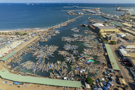 Aerial view of a bustling harbor filled with fishing boats their hulls reflecting the sunlight amid the darker waters contrasting with the sandy shore and port infrastructure Tema Ghana. Aerial view of a bustling harbor filled with fishing boats their hulls reflecting the sunlight amid the darker waters contrasting with the sandy shore and port infrastructure Tema Ghana.