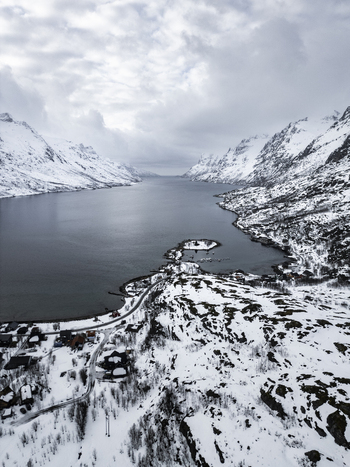Aerial view of Ersfjorden fjord surrounded by steep snow capped mountains and a small coastal village under a cloudy sky Ersfjord Troms Norway. Aerial view of Ersfjorden fjord surrounded by steep snow capped mountains and a small coastal village under a cloudy sky Ersfjord Troms Norway.