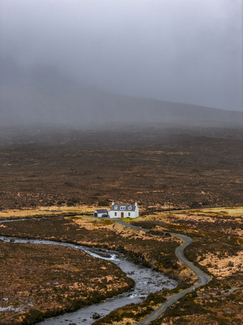 Aerial view of a lonely white cottage nestled in the rugged moorland landscape with a winding road and a stream under a misty sky Isle of Skye Scotland United Kingdom. Aerial view of a lonely white cottage nestled in the rugged moorland landscape with a winding road and a stream under a misty sky Isle of Skye Scotland United Kingdom.