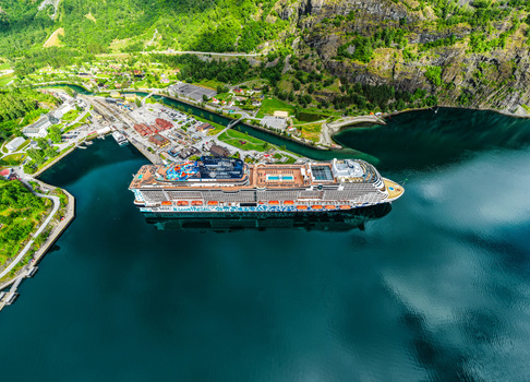 Aerial view of a large cruise ship docked in the deep blue waters of the Aurlandsfjord surrounded by lush green mountains and the small village of Flam Vestland Norway. Aerial view of a large cruise ship docked in the deep blue waters of the Aurlandsfjord surrounded by lush green mountains and the small village of Flam Vestland Norway.