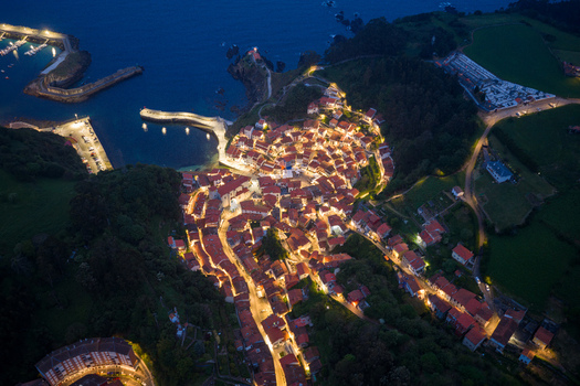 Aerial view of the illuminated coastal town of Cudillero at night with glowing streetlights red roofed houses and the harbor piers reflecting in the dark water Cudillero Asturias Spain. Aerial view of the illuminated coastal town of Cudillero at night with glowing streetlights red roofed houses and the harbor piers reflecting in the dark water Cudillero Asturias Spain.