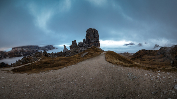 View of the Cinque Torri rock formation in the Dolomites with a gravel path leading through autumnal hills under a dramatic cloudy sky and low fog Trentino Alto Adige Italy. View of the Cinque Torri rock formation in the Dolomites with a gravel path leading through autumnal hills under a dramatic cloudy sky and low fog Trentino Alto Adige Italy.