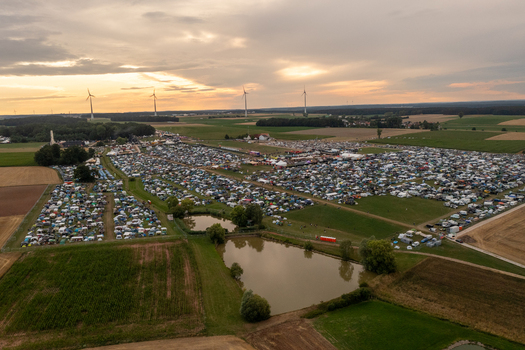 Aerial view of the Open Beatz Festival campsite with numerous tents a small pond and wind turbines on the horizon under a sunset sky in Herzogenaurach Bayern Germany. Aerial view of the Open Beatz Festival campsite with numerous tents a small pond and wind turbines on the horizon under a sunset sky in Herzogenaurach Bayern Germany.