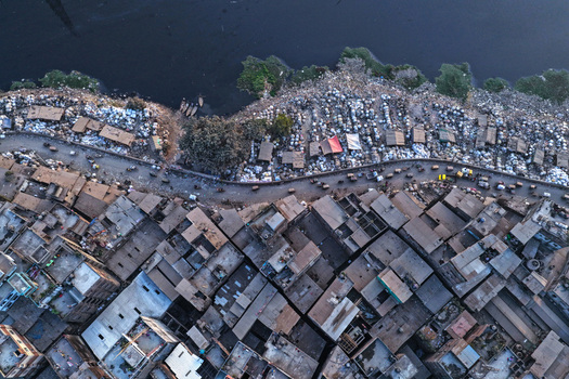 Aerial view of Buriganga River flowing past a dense urban neighborhood with crowded rooftops a busy road and riverbank activity in Dhaka Dhaka Division Bangladesh. Aerial view of Buriganga River flowing past a dense urban neighborhood with crowded rooftops a busy road and riverbank activity in Dhaka Dhaka Division Bangladesh.