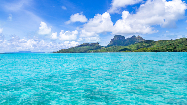View of the turquoise lagoon of Bora Bora with Mount Otemanu rising in the background under a bright blue sky with white clouds in Bora Bora Leeward Islands French Polynesia.