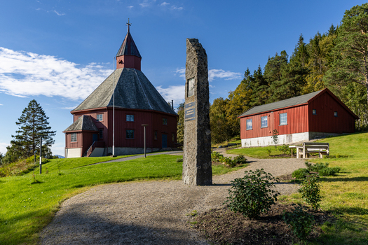 View of the red octagonal Hadsel Church and a stone monument on a green lawn surrounded by trees under a blue sky in Stokmarknes Nordland Norway. View of the red octagonal Hadsel Church and a stone monument on a green lawn surrounded by trees under a blue sky in Stokmarknes Nordland Norway.