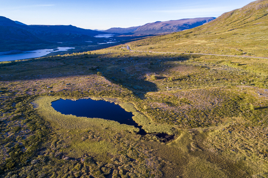 Aerial view of Jotunheimen mountain landscape with a small alpine lake grassy tundra rolling hills and a distant winding road in Jotunheimen Innlandet Norway.