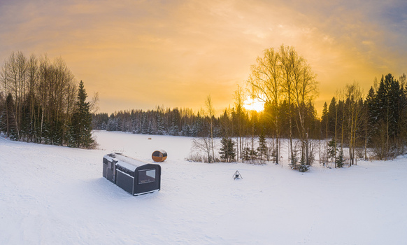 Aerial view of a modern cabin and a barrel sauna in a snowy landscape surrounded by pine trees during a golden sunset in Latvia. Aerial view of a modern cabin and a barrel sauna in a snowy landscape surrounded by pine trees during a golden sunset in Latvia.