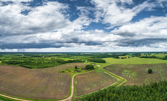 Aerial view of a rural farmstead surrounded by agricultural fields dirt roads and lush forests under a dramatic cloudy sky in Latvia. Aerial view of a rural farmstead surrounded by agricultural fields dirt roads and lush forests under a dramatic cloudy sky in Latvia.
