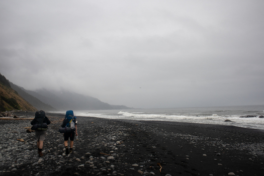 View of two hikers with large backpacks walking along the black sand beach with crashing waves and misty mountains at the Lost Coast in Humboldt County California United States.