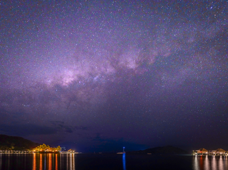 View of Likuliku Lagoon Resort with illuminated overwater bungalows reflecting on the calm ocean under a vibrant starry night sky with the Milky Way visible in Yaro Nadroga Navosa Fiji.