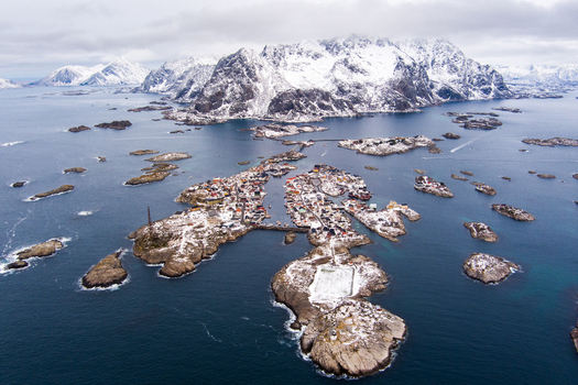 Aerial view of the village of Henningsvaer in the Lofoten archipelago featuring its iconic football pitch on a rocky island surrounded by snow capped mountains and the sea in Nordland Norway.