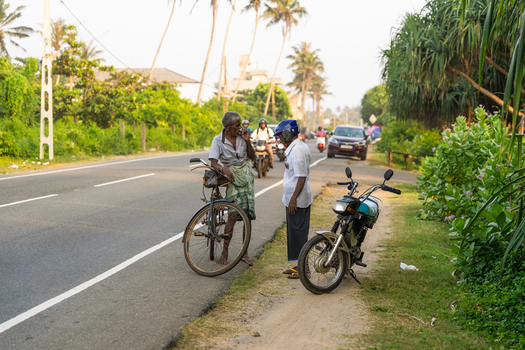 Sri Lanka 21 January 2026 View of two local men talking on the side of a coastal road with a bicycle and motorcycle under palm trees and bright daylight.