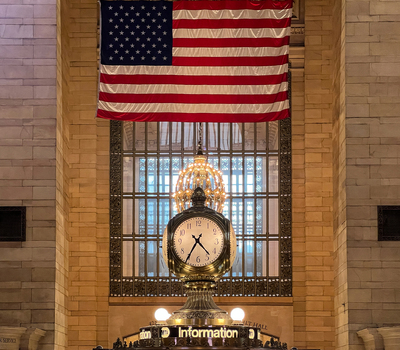 View of the iconic four faced clock at the information booth in Grand Central Terminal with a large American flag hanging above in New York New York United States. View of the iconic four faced clock at the information booth in Grand Central Terminal with a large American flag hanging above in New York New York United States.