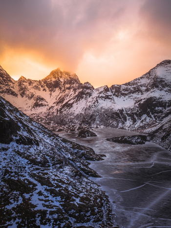 Aerial view of snow covered mountains embrace a frozen lake under a fiery sky Leknes Nordland Norway.
