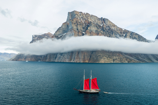 Aerial view of a sailboat with red sails in a fjord set against the dramatic backdrop of Storoen mountain partially shrouded in low hanging clouds in Avannaata Kommunia Greenland. Aerial view of a sailboat with red sails in a fjord set against the dramatic backdrop of Storoen mountain partially shrouded in low hanging clouds in Avannaata Kommunia Greenland.
