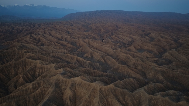 Aerial view of the rugged arid landscape of Issyk Kul with deep ridges undulating hills and distant snow capped mountains under a twilight sky in Issyk Kul Region Kyrgyzstan. Aerial view of the rugged arid landscape of Issyk Kul with deep ridges undulating hills and distant snow capped mountains under a twilight sky in Issyk Kul Region Kyrgyzstan.