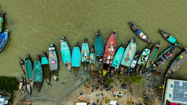 Aerial view of Baish Mowza Sarak Bazar with numerous colorful wooden boats docked along the riverbank and people gathering on the shore in Nabinagar Chittagong Division Bangladesh. Aerial view of Baish Mowza Sarak Bazar with numerous colorful wooden boats docked along the riverbank and people gathering on the shore in Nabinagar Chittagong Division Bangladesh.