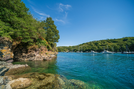 View of the Fowey River with moored boats rocky shoreline and lush green hills under a clear blue sky in Fowey Cornwall United Kingdom. View of the Fowey River with moored boats rocky shoreline and lush green hills under a clear blue sky in Fowey Cornwall United Kingdom.