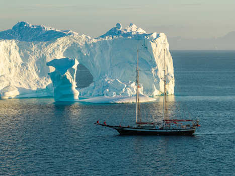 Aerial view of a sailboat navigating near a massive arch iceberg in the deep blue waters of Greenland. Aerial view of a sailboat navigating near a massive arch iceberg in the deep blue waters of Greenland.