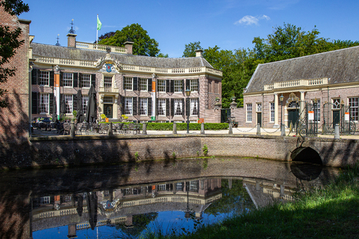 View of Castle Groeneveld with its historic brick facade reflecting in the moat water and a stone bridge under a clear blue sky in Baarn Utrecht Netherlands.