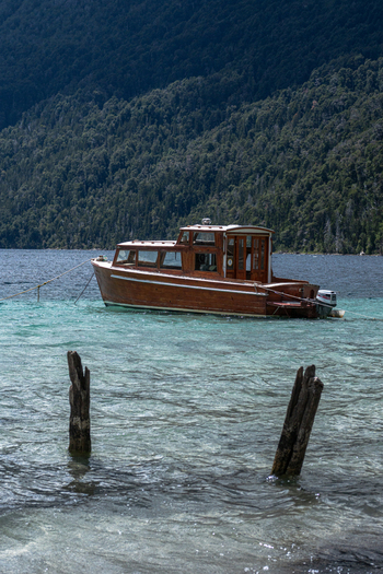 View of a wooden boat anchored in the turquoise waters of Bahia Brava with weathered posts in the foreground and a forested mountain in Villa La Angostura Neuquen Province Argentina. View of a wooden boat anchored in the turquoise waters of Bahia Brava with weathered posts in the foreground and a forested mountain in Villa La Angostura Neuquen Province Argentina.