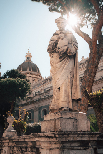 View of a marble statue bathed in sunlight standing proudly before a grand building with a dome amidst vibrant green trees Taormina Sicily Italy. View of a marble statue bathed in sunlight standing proudly before a grand building with a dome amidst vibrant green trees Taormina Sicily Italy.