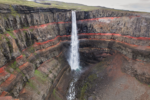 Aerial view of Hengifoss waterfall cascading down steep cliffs with visible red clay layers and basaltic rock formations in Egilsstadir Fljotsdalshreppur Iceland. Aerial view of Hengifoss waterfall cascading down steep cliffs with visible red clay layers and basaltic rock formations in Egilsstadir Fljotsdalshreppur Iceland.