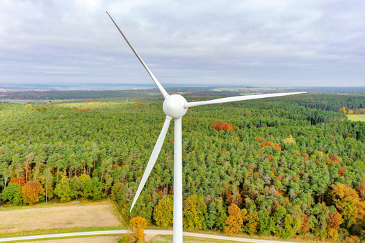 Aerial view of a large white wind turbine standing above a dense forest with vibrant autumn foliage under an overcast sky in Simmerath Nordrhein Westfalen Germany. Aerial view of a large white wind turbine standing above a dense forest with vibrant autumn foliage under an overcast sky in Simmerath Nordrhein Westfalen Germany.
