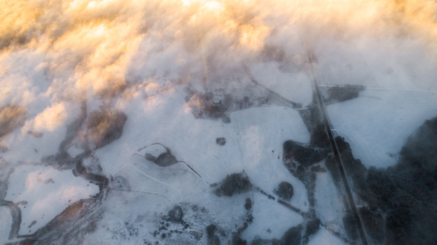 Aerial view of snow covered fields and a road partially obscured by low hanging clouds during golden hour in Latvia. Aerial view of snow covered fields and a road partially obscured by low hanging clouds during golden hour in Latvia.