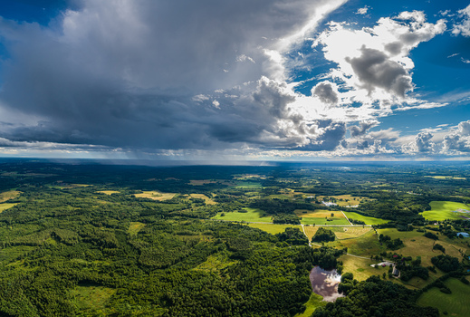 Aerial view of lush green forests agricultural fields and a small pond under a dramatic sky with heavy rain clouds and bright sunlight in Latvia. Aerial view of lush green forests agricultural fields and a small pond under a dramatic sky with heavy rain clouds and bright sunlight in Latvia.