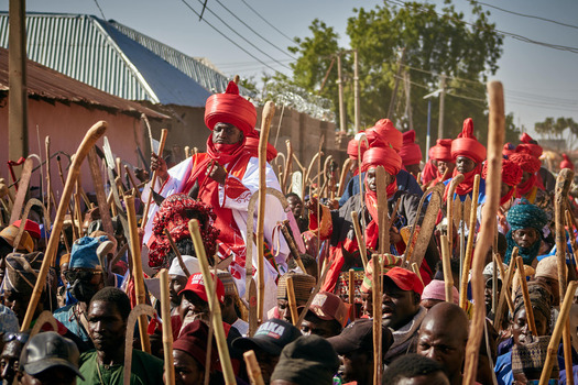 Zaria Nigeria 21 March 2026 View of a vibrant procession of figures in bold red and white holding rustic wooden staffs under a bright sky. Zaria Nigeria 21 March 2026 View of a vibrant procession of figures in bold red and white holding rustic wooden staffs under a bright sky.