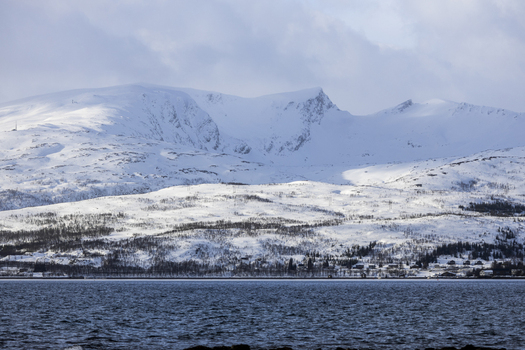 View of a snow covered mountain range and a calm fjord under a cloudy sky with small houses along the coastline in Kvaloya Troms Norway. View of a snow covered mountain range and a calm fjord under a cloudy sky with small houses along the coastline in Kvaloya Troms Norway.