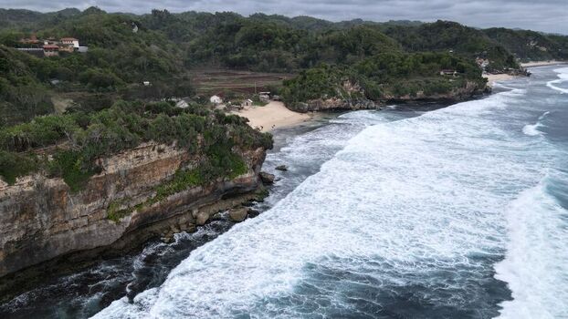 Aerial view of Indrayanti Beach with rugged limestone cliffs white sand crashing waves and lush green vegetation in Kecamatan Tepus Daerah Istimewa Yogya Indonesia. Aerial view of Indrayanti Beach with rugged limestone cliffs white sand crashing waves and lush green vegetation in Kecamatan Tepus Daerah Istimewa Yogya Indonesia.