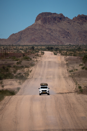 View of a white vehicle kicking up dust journeying down a long straight desert road toward a distant mountain under a clear blue sky Keetmanshoop Karas Region Namibia. View of a white vehicle kicking up dust journeying down a long straight desert road toward a distant mountain under a clear blue sky Keetmanshoop Karas Region Namibia.