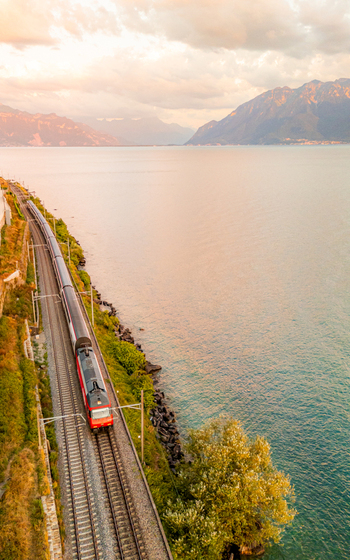 Aerial view of a red train traveling along the tracks beside Lake Geneva during golden hour in Bourg en Lavaux Vaud Switzerland. Aerial view of a red train traveling along the tracks beside Lake Geneva during golden hour in Bourg en Lavaux Vaud Switzerland.