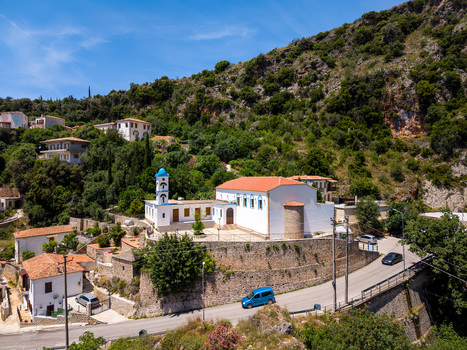 Aerial view of a traditional white church with a blue dome and red tiled roof nestled on a green hillside with stone walls and a winding road in Dhermi Vlore County Albania.