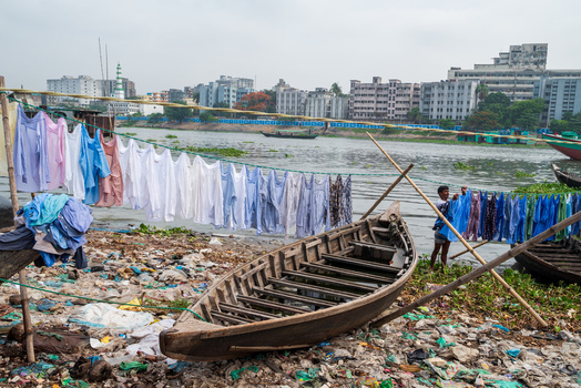 Dhaka Division Bangladesh 14 May 2026 View of the Buriganga River with a man hanging laundry on lines next to a wooden boat on a polluted bank.